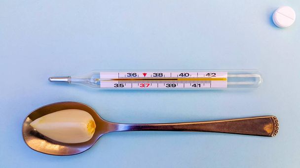 on a light blue background, a thermometer showing a high temperature, a spoon with garlic, and in the corner, a tablet, all this symbolizes the right choice of treatment. - Photo, Image
