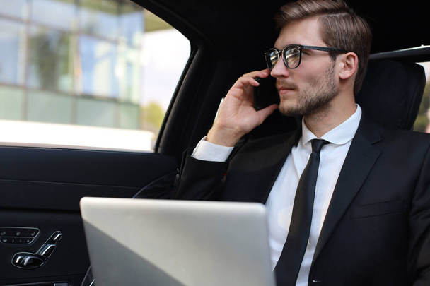 Όμορφος επιχειρηματίας με αυτοπεποίθηση που μιλάει στο έξυπνο τηλέφωνο και εργάζεται χρησιμοποιώντας φορητό υπολογιστή ενώ κάθεται στο αυτοκίνητο. - Φωτογραφία, εικόνα