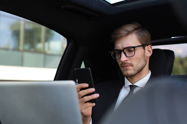 Handsome confident man in full suit looking at his smart phone while sitting in the car and using laptop. - Foto, imagen
