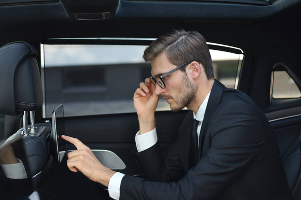 Thoughtful young businessman sitting in the luxe car and using his tablet. - Foto, afbeelding