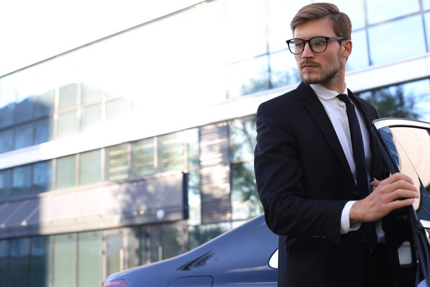 Handsome serious young businessman standing near his comfortable new car and going to the office. - Foto, Imagem