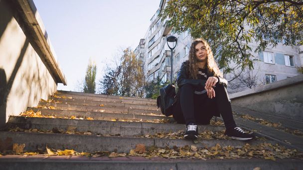 A young beautiful woman in casual clothes is sitting on the steps with leaves in the urban district. - Photo, Image