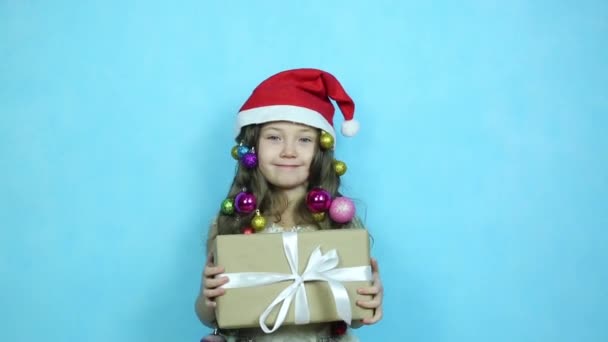 Κοριτσάκι σε ένα όμορφο φόρεμα με μπάλες Χριστουγέννων στα μαλλιά της κρατά ένα κουτί έκπληξη δώρο για τα Χριστούγεννα στα χέρια της. - Πλάνα, βίντεο