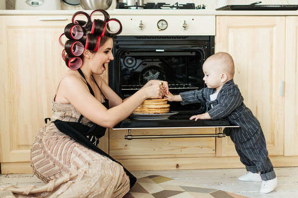 A young mother in an apron and curlers is preparing a birthday cake in the kitchen with her little child - Photo, Image