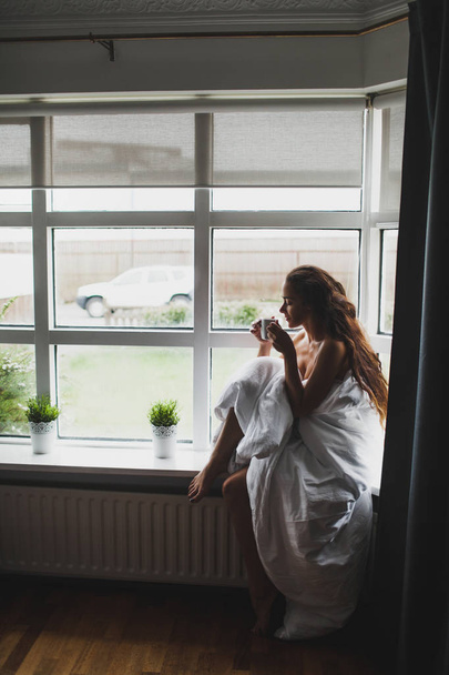 Yung mujer cubierta con manta sentado en el alféizar de la ventana y disfrutar
 - Foto, imagen