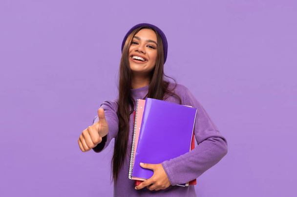 Hymyilevä afrikkalais-amerikkalainen tto teini-ikäinen opiskelija hattu ja villapaita tilalla kansiot ja osoittaa peukalo pystyssä eristetty violetti tausta
. - Valokuva, kuva