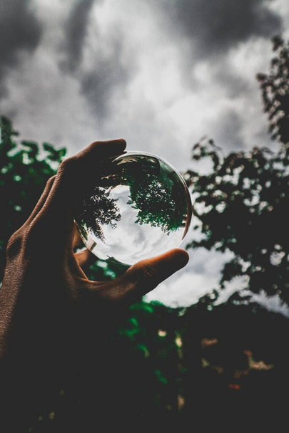 A vertical selective closeup shot of a person holding a crystal ball with the reflection of beautiful green trees and the breathtaking clouds - Φωτογραφία, εικόνα
