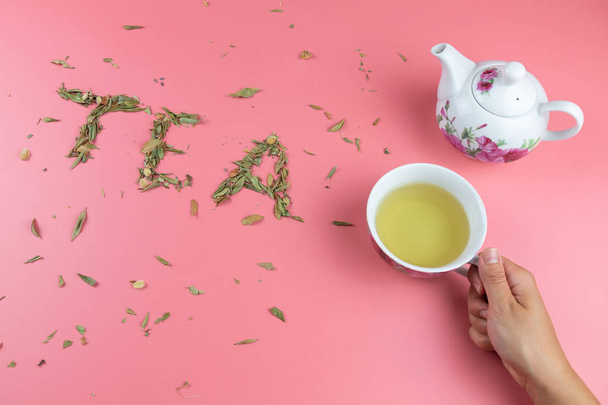 γυναικείο χέρι κρατώντας ένα φλιτζάνι τσάι με μια λευκή τσαγιέρα στο πλάι και οι λέξεις τσάι γραμμένο με χαλαρό τσάι φύλλο σε ένα φωτεινό ροζ φόντο - Φωτογραφία, εικόνα