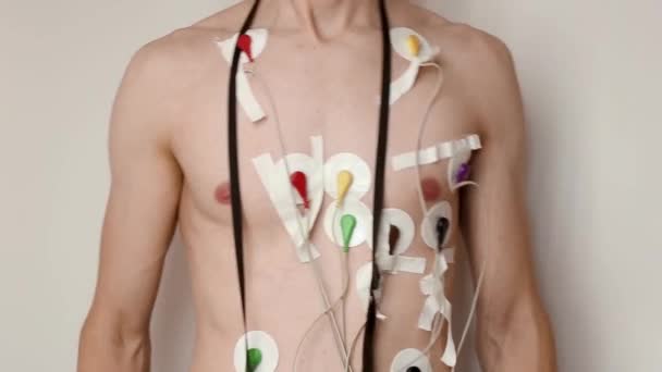 Beyaz arka planda göğsüne bağlı Holter monitörü olan genç, zayıf bir adam. Kablolar yamayla cilde bağlanmış. 24 saat boyunca elektriksel kardiyogram kaydı, sağlık ve kalp sorunları teşhisi - Video, Çekim