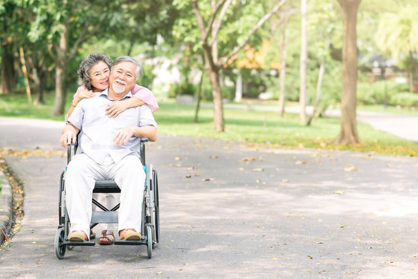 ηλικιωμένη γυναίκα αγκαλιάζει τον άντρα της σε αναπηρική καρέκλα από πίσω  - Φωτογραφία, εικόνα