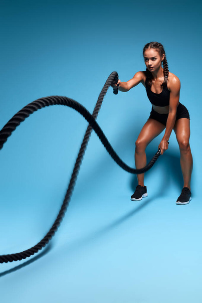 An attractive young muscular motivated athletic girl using battle ropes at gym. - Фото, изображение