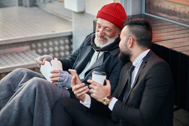 Young positive businessman and mature beggar look at phone - Foto, Bild