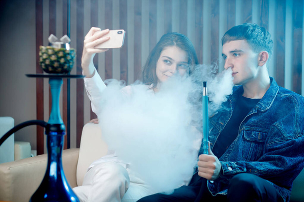 Beautiful young woman with guy hookah, blow smoke from tobacco and take selfie photo on phone, blue shisha background - Photo, Image