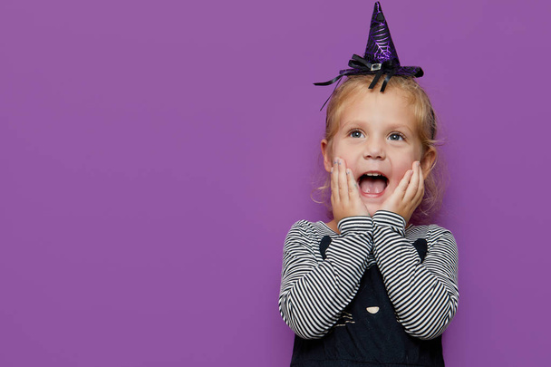 Happy Halloween! Cute shocked little child girl in witch costume and hat on violet or purple background.  - Фото, зображення