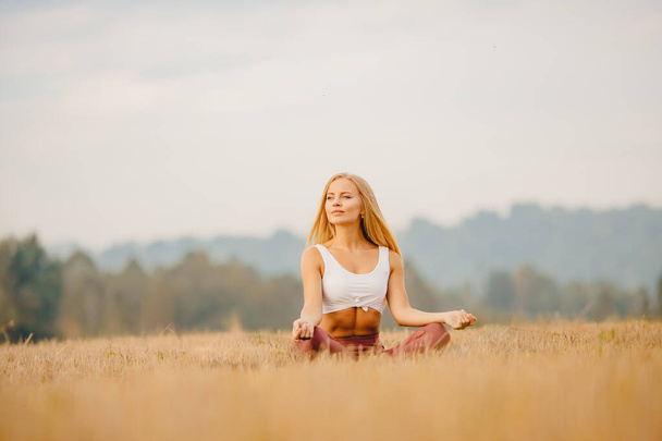 Giovane ragazza in camicia bianca, abbigliamento sportivo con i capelli lunghi Bionda medita yoga in campo alba. Concetto di relax
 - Foto, immagini