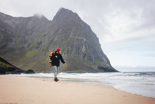 Alone tourist walking along the sea beach among foggy mountains. Adventure traveler with backpack hiking by ocean sand - Фото, зображення