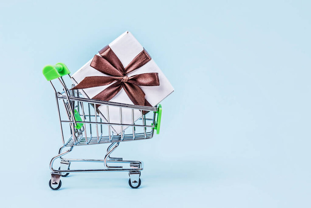 Gift box with brown bow in miniature shopping cart on pastel blue background with copy space. - Foto, afbeelding