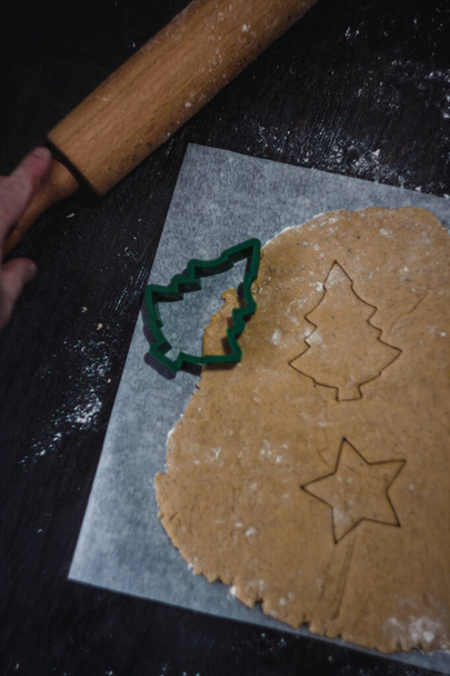 вырезать пряничное печенье в виде рождественской елки из сырого теста на бумаге для выпечки пергамента на темном фоне. Вид сверху. сохранить место
 - Фото, изображение