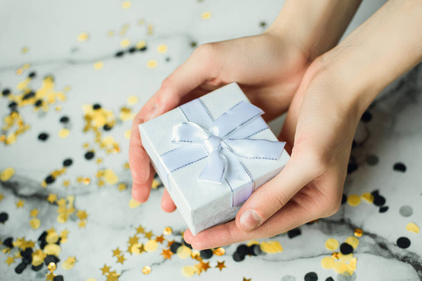 Серебряный подарок в руках девушки без маникюра на сером фоне с золотыми и черными конфетти. Концепция Нового года и Рождества. Вид сбоку
 - Фото, изображение