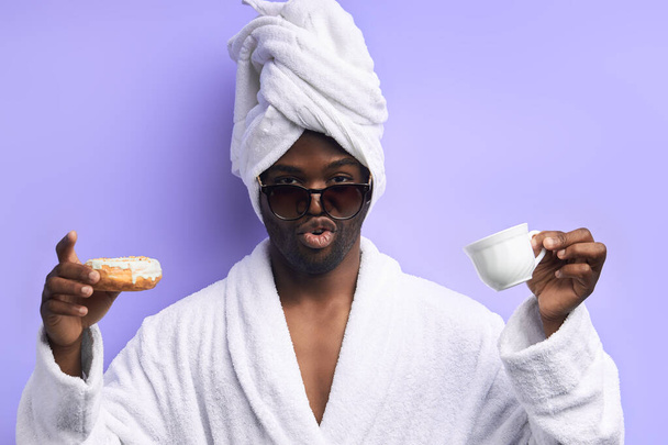 Excited young african man holding glazed donut and cup of tea or coffee - Fotó, kép