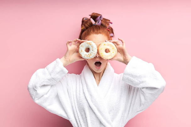 Portrait of young redhead girl in bathrobe with curlers on hair, posing with donuts - Фото, изображение