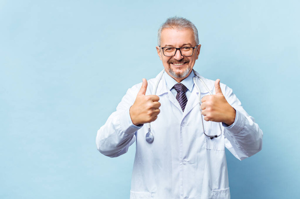 Uśmiechnięty starszy lekarz ze stetoskopem. Na niebieskim tle. Sanitariusz pokazuje podniesiony kciuk. Koncepcja zwycięstwa ludzkości nad chorobą - Zdjęcie, obraz