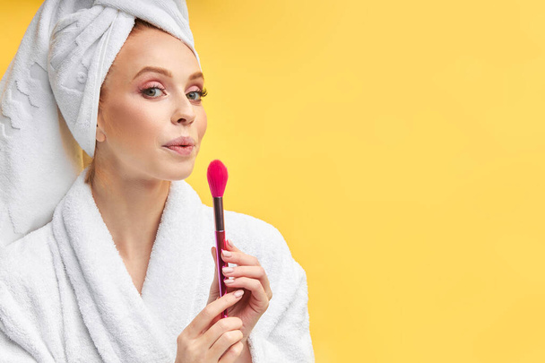 Retrato de mulher bonita soprando escova de maquiagem usando roupão de banho e toalha
 - Foto, Imagem