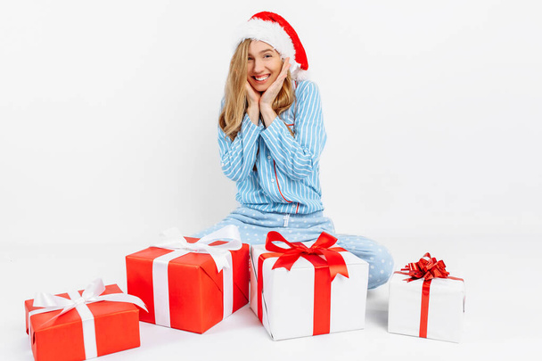 Christmas morning. Happy beautiful young girl, in Christmas pajamas and Santa hat, received many Christmas gifts - Фото, изображение