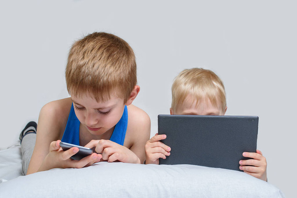 ガジェットを持った二人の少年がベッドに横たわっている。子供はスマートフォンとタブレットを使用します - 写真・画像