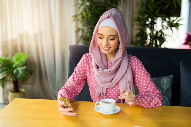 beautiful young woman in muslim dress is drinking tea in a restaurant with a phone in her hand - Foto, Imagen