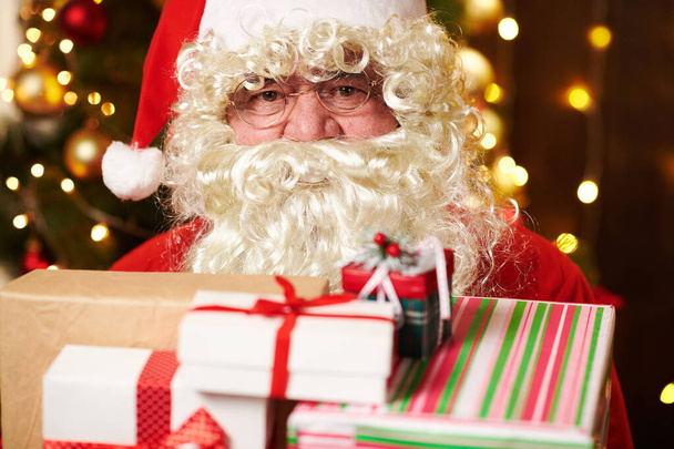 Kerstman poseren met vele geschenken, binnen zitten in de buurt van versierde kerstboom met verlichting - Vrolijk kerstfeest en fijne feestdagen! - Foto, afbeelding