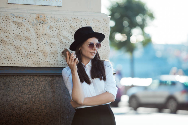 young woman in a hat and a white shirt walks in the city and uses a smartphone. - Foto, immagini
