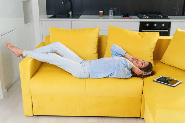 Pretty blonde teenage girl in headphones is listening to music, looking at camera and smiling while lying on her back, legs up, on sofa at home - Foto, imagen
