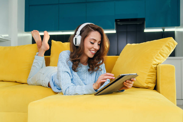 Exuberant girl uses tablet pc while laying on the comfortable yellow sofa and enjoying nice melodies with earphones. - Φωτογραφία, εικόνα