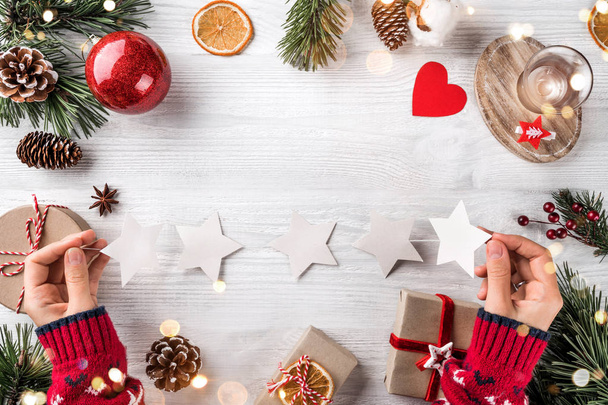 Női kéz kezében egy koszorú csillagokkal karácsonyi háttérben faágakkal, fenyőtobozokkal, ajándékokkal. Karácsony és Boldog Új Évet, bokeh, fény. Lapos fekvésű, felülnézet - Fotó, kép
