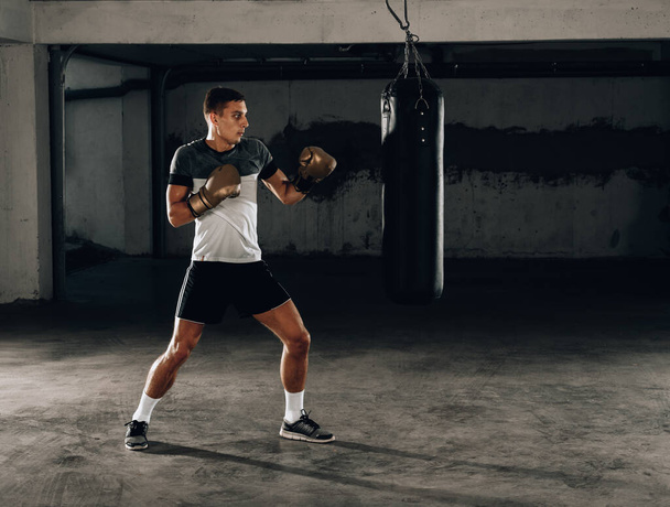 muskulöser, gutaussehender Kämpfer, der während einer Trainingsrunde mit einer Boxtasche einen kraftvollen Tritt nach vorne gibt - Foto, Bild