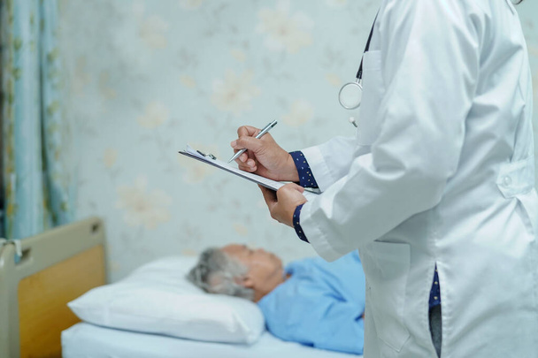 Врач записывает диагноз на планшет, в то время как пожилая женщина азиатского возраста или пожилая женщина лежит на кровати в палате медсестер: здоровый сильный медицинский концепт - Фото, изображение
