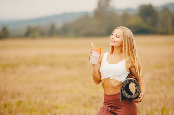 Mooi blond meisje lessen en drinkt dorst met fles water, yoga mat, in achtergrond park - Foto, afbeelding
