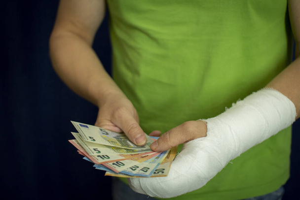 Ένας άντρας με ένα χέρι σε γύψο κρατάει χρήματα από το κόστος και τα έξοδα της επείγουσας ιατρικής περίθαλψης σε ένα νοσοκομείο. . - Φωτογραφία, εικόνα