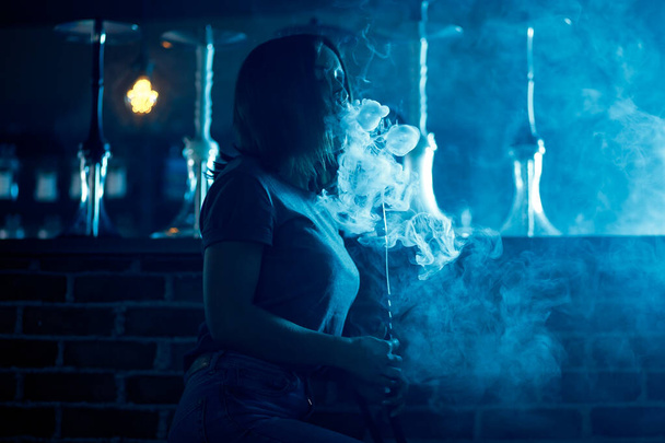 Beautiful woman blows smoke rings in night club hookah or shisha bar, blue background - Foto, imagen