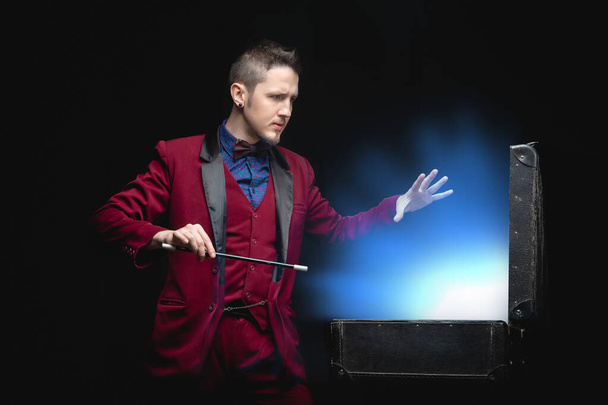 Zauberer Mann mit Zauberstab zeigt Verschwinden im Koffer blaues Glühlicht auf schwarzem Hintergrund - Foto, Bild
