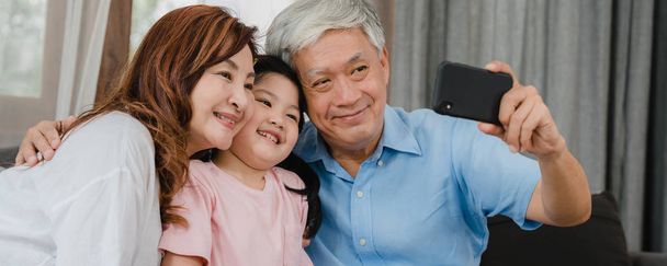 asiatische Großeltern Selfie mit Enkelin zu Hause. Senior chinesische glücklich verbringen Familienzeit mit Handy entspannen mit jungen Mädchen Kind auf dem Sofa im Wohnzimmer liegend. panoramische Banner Hintergrund. - Foto, Bild