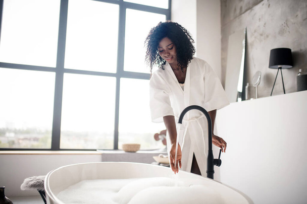 Curly dark-skinned woman preparing bath with foam - Fotó, kép