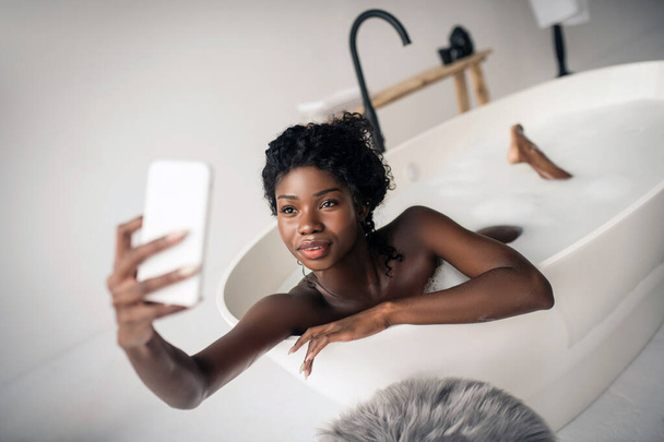 Dark-eyed woman making selfie while having foamy bath - Fotó, kép