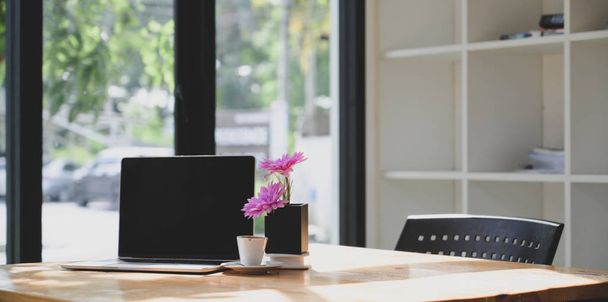 Ordenador portátil de pantalla en blanco abierto en un cómodo espacio de trabajo con flor rosa en maceta de árbol
  - Foto, Imagen