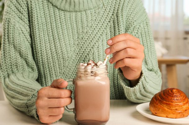 Woman holding mason jar of delicious cocoa drink with marshmallows at white table, closeup - Foto, Imagen