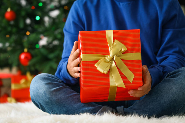 Man in blue sweater with Christmas gift sitting on fluffy carpet at home, closeup - Foto, immagini