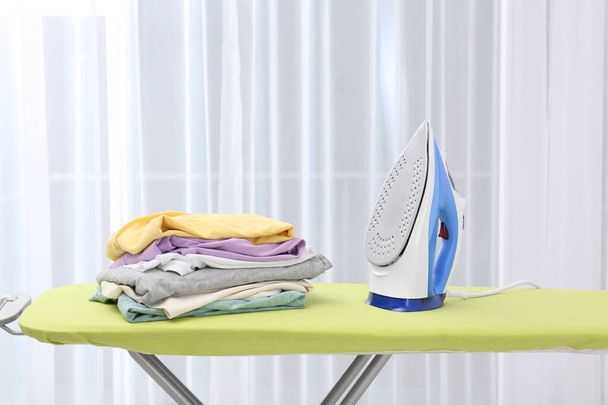 Board with modern iron and stack of clothes indoors. Laundry day - Photo, image