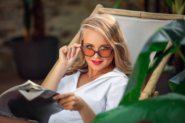 Όμορφη νεαρή γυναίκα ανάγνωση περιοδικών ενώ χαλαρώνετε στην ξαπλώστρα στο resort - Φωτογραφία, εικόνα