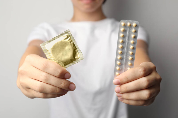 Young woman holding birth control pills and condom on light grey background, closeup. Safe sex concept - 写真・画像